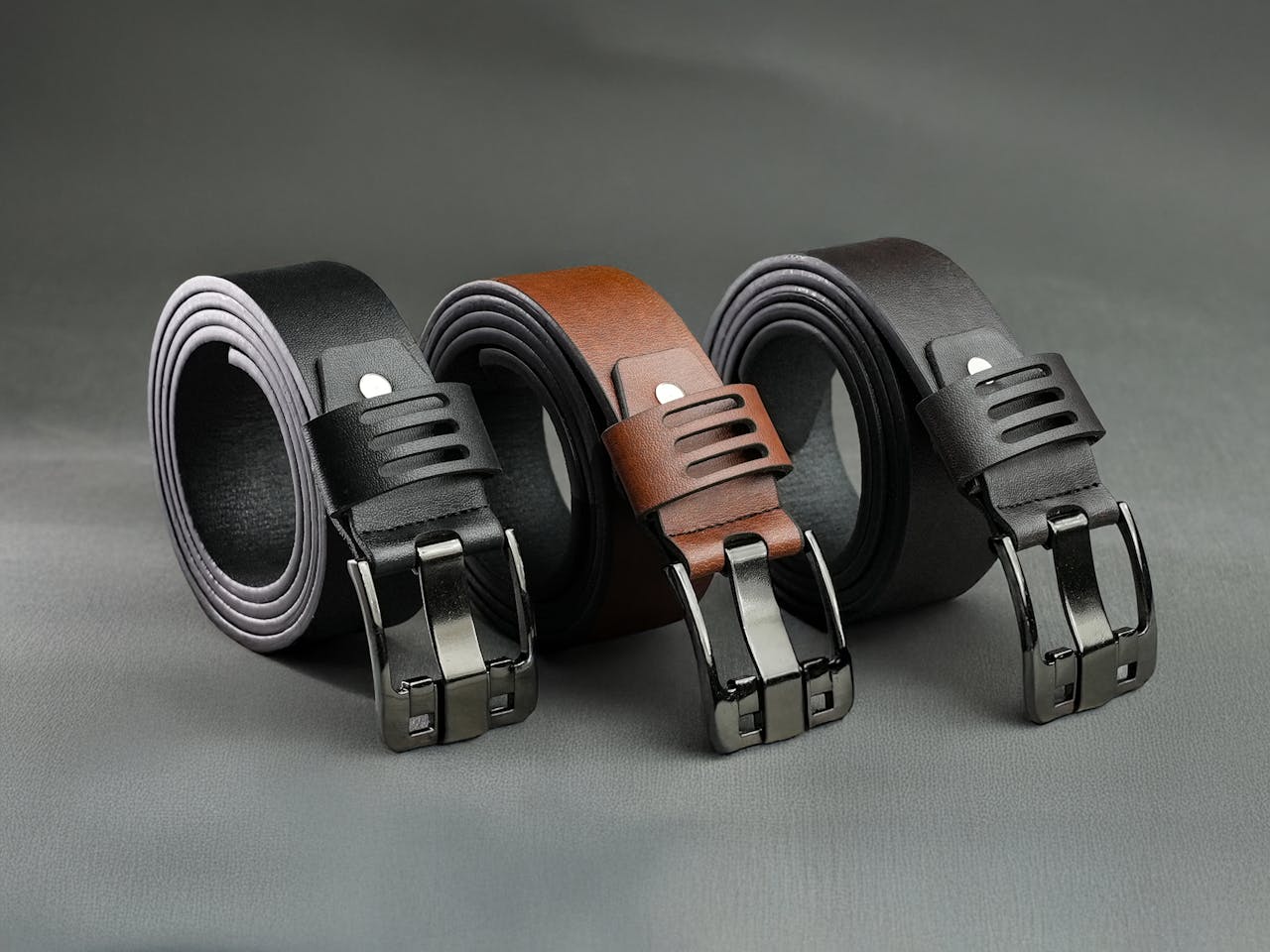 Three stylish leather belts with modern buckles on a gray background.